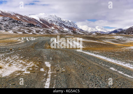 A picturesque autumn landscape with a rocky dirt road through the steppe in the mountains, golden trees on the slopes and the first snow Stock Photo