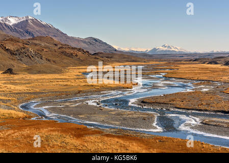 A beautiful autumn landscape with a winding river in the valley between the mountains, ice on the banks, golden grass and mountains covered with snow  Stock Photo