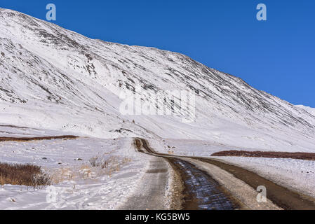 Picturesque view with snowy mountain peaks, a gravel road through the pass high in the mountains and the first snow on the background of the blue sky Stock Photo