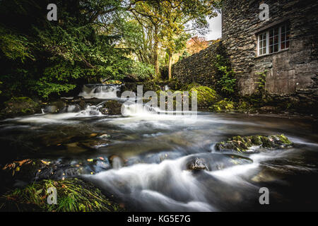 A river flows over rocks and past a mill on its way into Coniston in the Lake District National Park Stock Photo