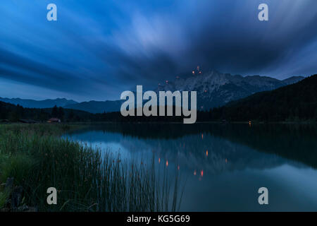 St. John's fire and rockets at the western side of Karwendel (mountain) (mountain) over Mittenwald. in foreground the idyllic Lautersee (lake) with reed and forest, over that move cloud veils resulted by long time exposure Stock Photo