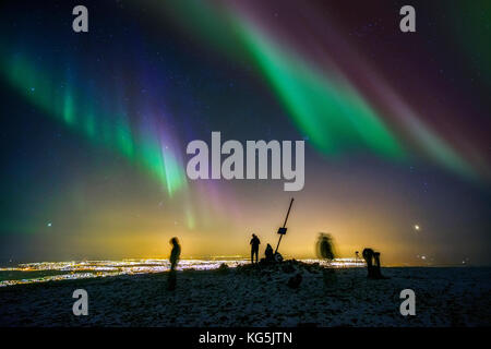 People enjoying the Aurora Borealis or Northern lights on Mt. Ulfarsfell, near Reykjavik, Iceland. The Hellisheidi Power Plant is off in the distance. Stock Photo