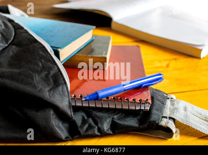 Open School black backpack, or bag, with three books and notebooks and blue pen spilling slight out of it, on top of a yellow wooden table surface Stock Photo