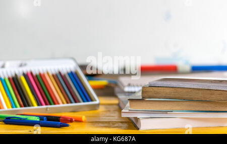 Coloring pencils, pens and closed books and notebooks on top of a yellow wooden table, with a white school board in the background Stock Photo
