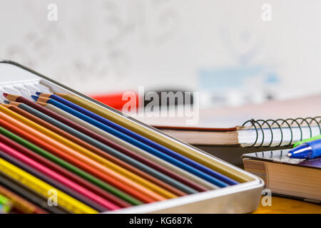 Coloring pencils, pens and closed books and notebooks on top of a yellow wooden table, with a white school board in the background Stock Photo