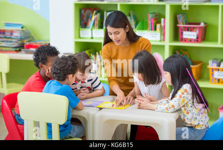 Asian female teacher teaching mixed race kids reading book in classroom,Kindergarten pre school concept Stock Photo