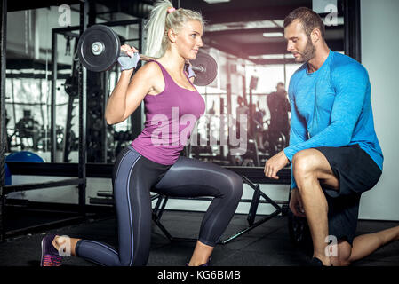 Young attractive blonde female doing lunge exercise in modern fitness center with assistance of her personal trainer. Stock Photo