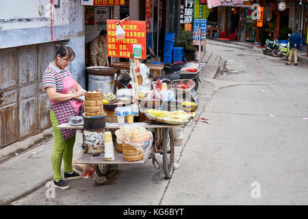 Lijiang, Yunnan, China - September 27, 2017: Woman prepares and sells breakfast food from street stall. Payment via QR code becomes very common in Chi Stock Photo