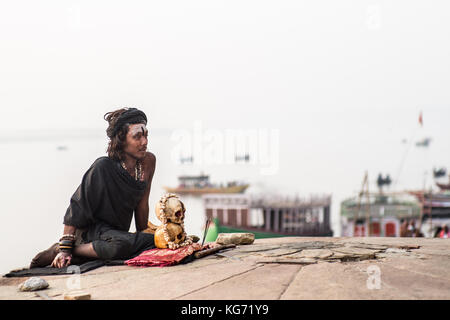 An Agori-yogi (hindu devotee) sitting with his human skulls next to the holy Ganges river and praying in the sunrise. Stock Photo