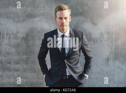 Stylish young businessman standing at the gray wall putting the hands in pockets. Stock Photo