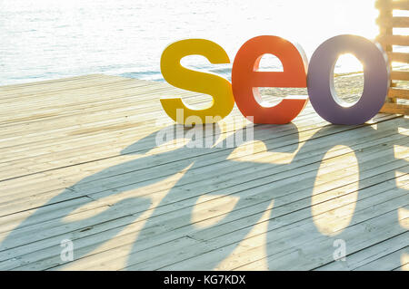 The word SEO by large colored letters creating a shadow on the wooden terrace on the river bank. Sun backlight. Selective focus. Space for text. Stock Photo