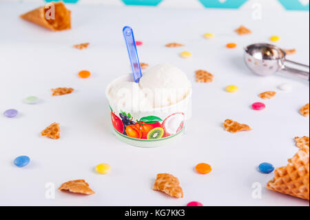 Close up Vanilla Ice-cream in a paper cup on the white background with colorful candies, pieces of waffles, ice cream spoon and empty ice cream cone.  Stock Photo