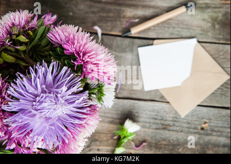 Top view Multicolor aster flowers bouquet, pen, blank white greeting card and craft paper envelope on rustic wooden table.. Postcard mock up. Autumn f Stock Photo
