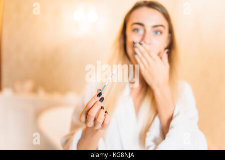 Frightened young woman in white bathrobe with positive pregnancy test, bathroom interior on background. Bodycare and hygiene, healthcare Stock Photo