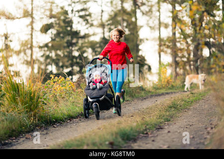 Running mother with child in stroller enjoying motherhood at autumn sunset and mountains landscape. Jogging or power walking woman with pram in woods. Stock Photo