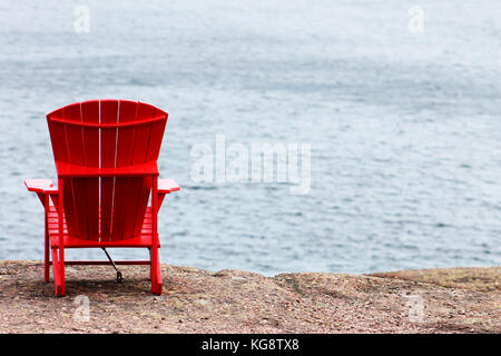 A red Adirondack chair anchored to the cliff, facing out toward the sea, trail side, Signal Hill, St. John's, Newfoundland and Labrador. Stock Photo