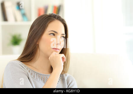 Portrait of a pensive woman wondering looking through a window sitting on a sofa in the living room at home with copy space in white Stock Photo