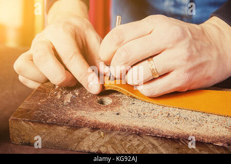 A close-up of a man skinner makes holes in the brown belt of full-length leather for fasteners on the wrinkled desk in the workshop. Saddler  manufact Stock Photo