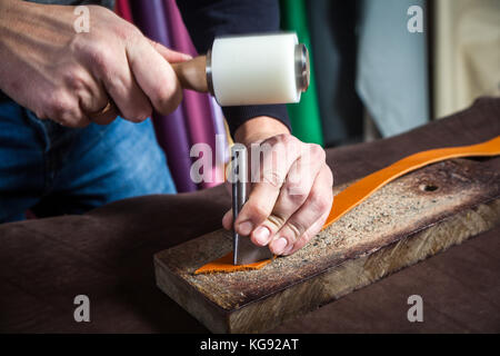 A close-up of a man skinner makes holes  with the help of a special hammer in the brown belt of full-length leather for fasteners on the wrinkled desk Stock Photo