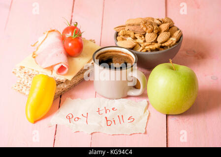 The message "pay the bills" wrote on a piece of paper placed on a pink wooden table, near a small cup of coffee and a morning meal Stock Photo