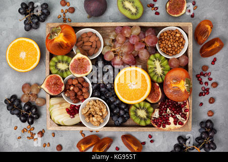 wooden tray with fresh fruits and nuts: orange, kiwi, figs, persimmon, grapes, almonds, walnuts, pine nuts, hazelnuts on a gray concrete background. v Stock Photo