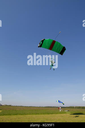 This skydiver is flying under his canopy in the sky. Soon he is going to land his green and black parachute on the grass area. Stock Photo