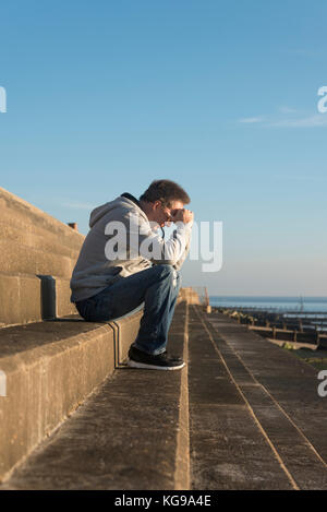 man with his head in his hands sitting on steps outside by the sea Stock Photo