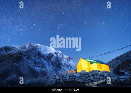 Yellow glowing tent against high rocks with snowy peak and sky with stars at night in Nepal. The Himalayan Mountains. Landscape with mountains, starry Stock Photo