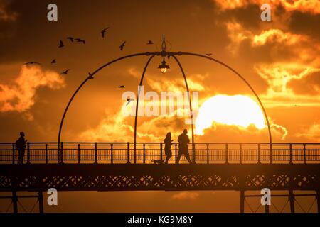 Southport, Merseyside, Sunset over Southport Pier. 5th November 2017. UK Weather.  People take a stroll along the boardwalk as a stunning sunset nestles into the horizon over the famous pier at Southport in Merseyside.  First opened in 1860, it spans a length of 1,108 metres (3,635 ft) and is the second longest in Great Britain. It was listed at Grade II on 18 August 1975. The  Tramway ran from the Promenade to the pier head at various times in the pier's history, most recently from August 2005 until June 2015.  Credit: Cernan Elias/Alamy Live News Stock Photo