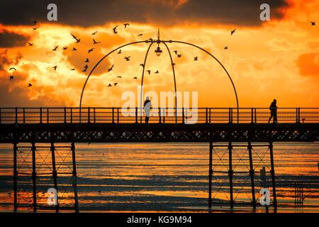 Southport, Merseyside, Sunset over Southport Pier. 5th November 2017. UK Weather.  People take a stroll along the boardwalk as a stunning sunset nestles into the horizon over the famous pier at Southport in Merseyside.  First opened in 1860, it spans a length of 1,108 metres (3,635 ft) and is the second longest in Great Britain. It was listed at Grade II on 18 August 1975. The  Tramway ran from the Promenade to the pier head at various times in the pier's history, most recently from August 2005 until June 2015.  Credit: Cernan Elias/Alamy Live News Stock Photo