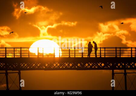 Southport, Merseyside, Sunset over Southport Pier. 5th November 2017. UK Weather.  People take a stroll along the boardwalk as a stunning sunset nestles into the horizon over the famous pier at Southport in Merseyside.  First opened in 1860, it spans a length of 1,108 metres (3,635 ft) and is the second longest in Great Britain. It was listed at Grade II on 18 August 1975. The  Tramway ran from the Promenade to the pier head at various times in the pier's history, most recently from August 2005 until June 2015.  Credit: Cernan Elias/Alamy Live News Stock Photo