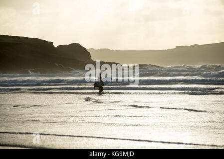 Strong winds and cold weather on the North Cornwall coast in bright autumn sunshine at Polzeath, Cornwall, UK. A surfer braves the cold and prepares to tackle the waves. Stock Photo