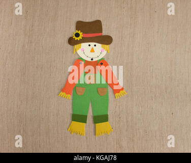 A felt scarecrow wearing orange, yellow, bright green and brown on a textured tan fabric background. Image has copy space. Stock Photo