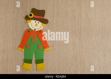 Felt scarecrow with tilted head wearing orange, yellow, bright green and brown on a textured tan fabric background. Image has copy space. Stock Photo