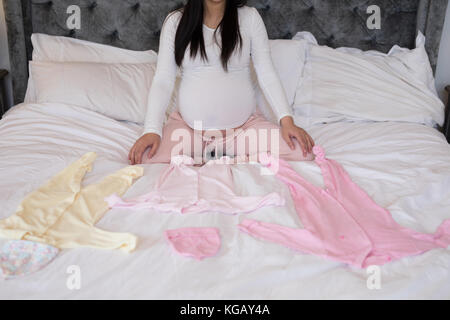 Mid section of pregnant woman sitting with baby clothes on bed Stock Photo