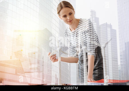 Professional attitude. Pleasant female engineer standing in the office and holding papers while looking on the windmill models Stock Photo