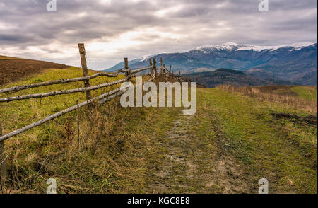 fence along dirt road in mountainous rural area. agricultural fields on hills in late autumn. mountain ridge with snowy tops in the distance Stock Photo