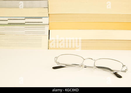 Pile of books novels on a white reflective surface with some silver glasses (spectacles) lying in the foreground. reading literacy concept Stock Photo
