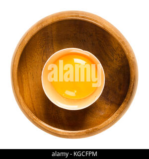 Open raw chicken egg with yolk and white in its shell in a wooden bowl. Common food and versatile ingredient used in cooking. Photo. Stock Photo