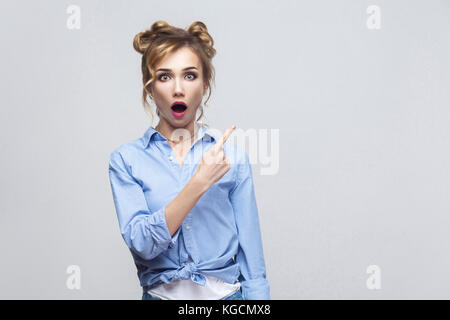 Excited blonde woman pointing finger sideways, raising eyebrows and keeping mouth wide opened, showing something surprising on grey copy space wall. I Stock Photo