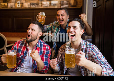 Men fans watching football on TV and drink beer. Three men drinking beer and having fun together in the bar Stock Photo