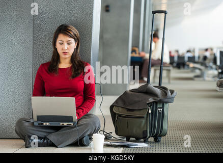 A businesswoman sitting on the floor at her departure gate at an airport working on her computer. Stock Photo