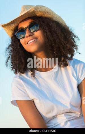 Beautiful happy mixed race African American female girl teenager young woman wearing aviator sunglasses and cowboy hat outside at golden sunset