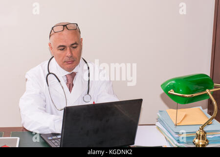 Doctor in his studio at desk, on pc,  with glasses. Use new technologies. In his professional studio, he is sitting at antique desk, and a green lamp  Stock Photo