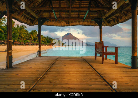 Wonderful view across the pier, on the left the tropical beach and in the background a beautiful mountain illuminated by red during sunset, Mauritius Stock Photo