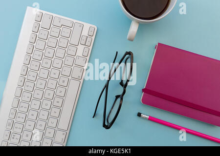 Computer keyboard eyeglasses and pink notebook overhead on blue background Stock Photo