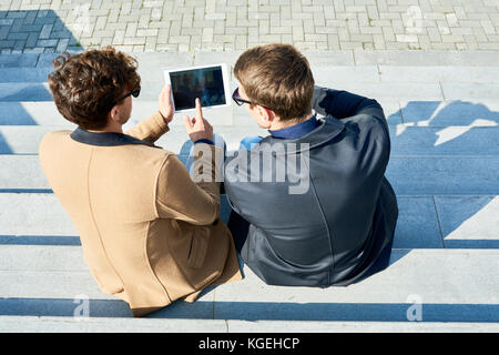 Back view  portrait of two handsome young men using digital tablet outdoors sitting on stone steps in city street Stock Photo