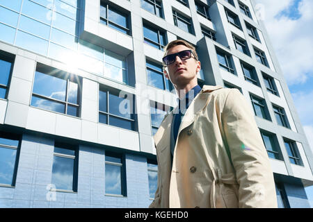 Low angle portrait of handsome young businessman wearing sunglasses and stylish trench coat standing outdoors looking away confidently against glass f Stock Photo