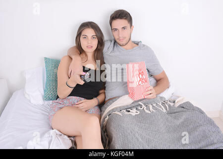 Young couple watching movies in bed and eating popcorn. Indoors. Stock Photo