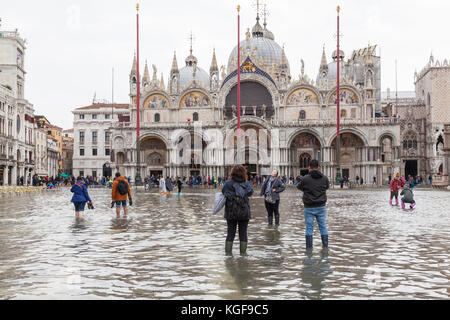 Venice, Veneto, Italy. 7th Nov, 2017. Acqua Alta high tide of 115cm from the lagoon causing temporary flooding in Piazza San Marco. Passarelle, or elevated walkways, are installed for pedestrian traffic. Happy woman posing in the water for her friends in front of Basilica San Marco. Water visible throughtout the Piazza San marco. Credit: Mary Clarke/Alamy Live News Stock Photo